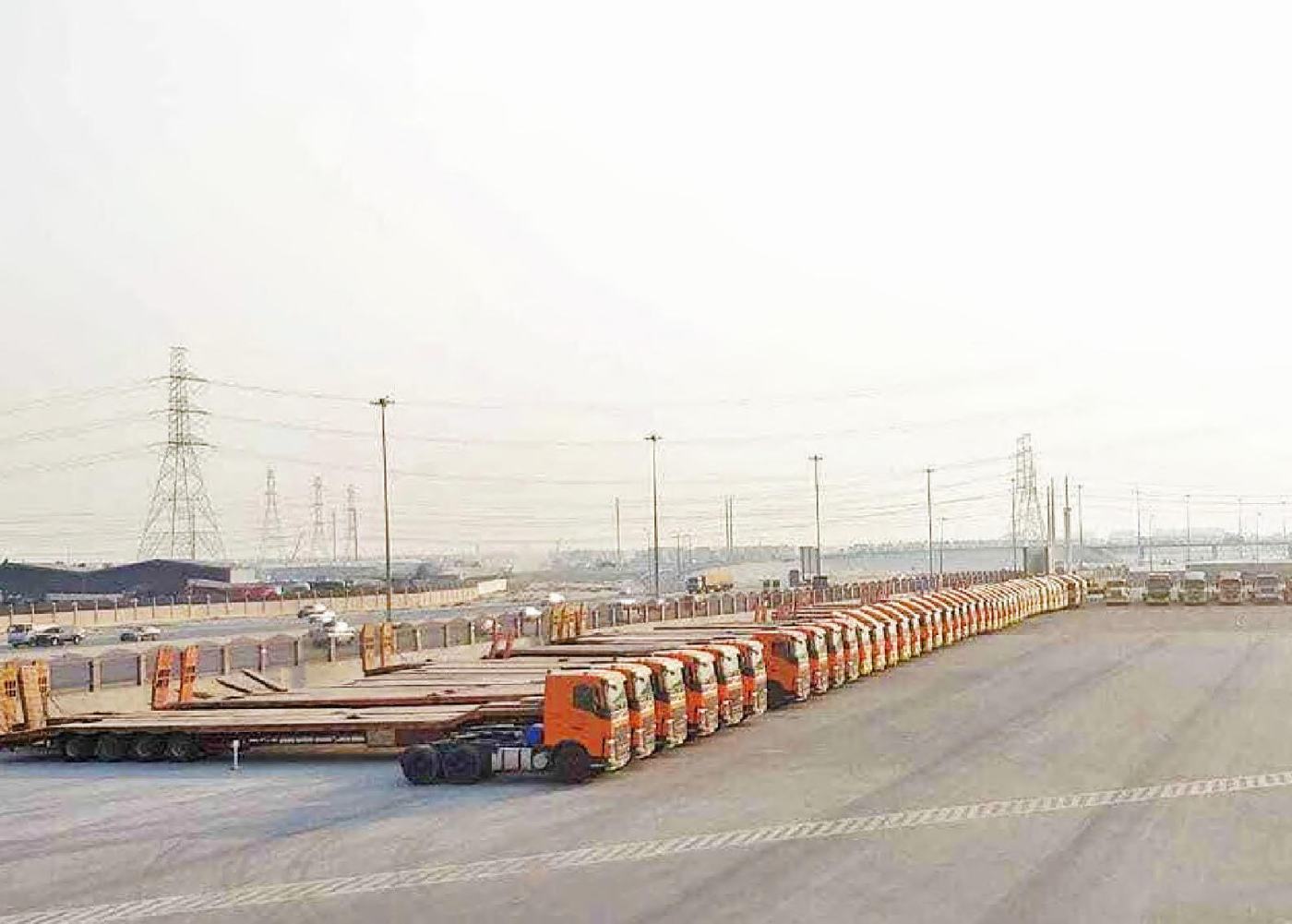 A long line of orange prime movers with lowbed trailers parked on a paved yard