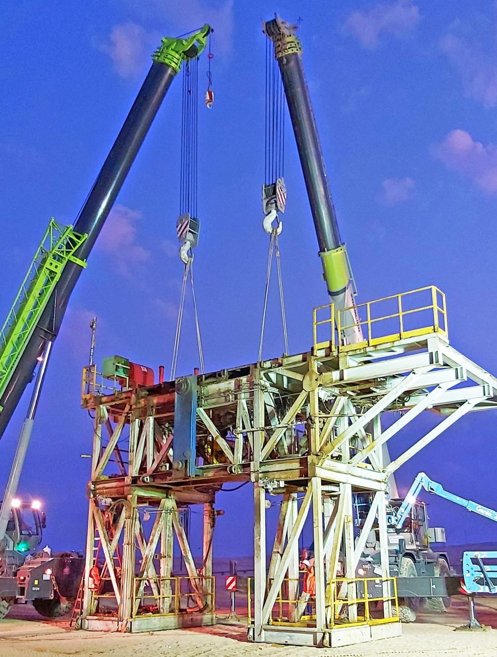 Two cranes lifting a rig substructure at dusk with floodlights on